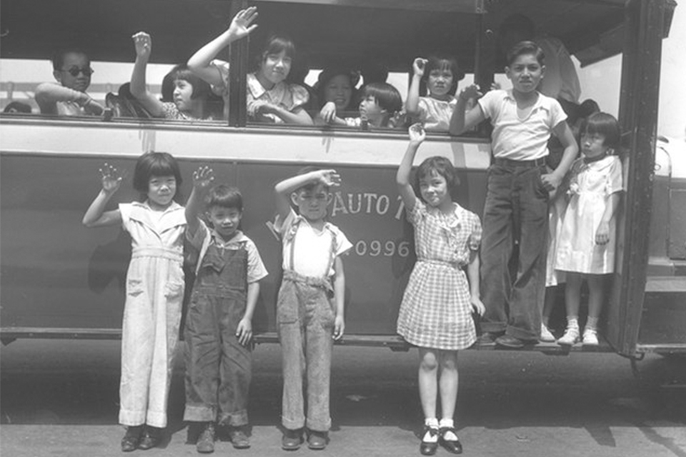 Black and white image of four children with raised left hands stand in front of a bus with many other children on it