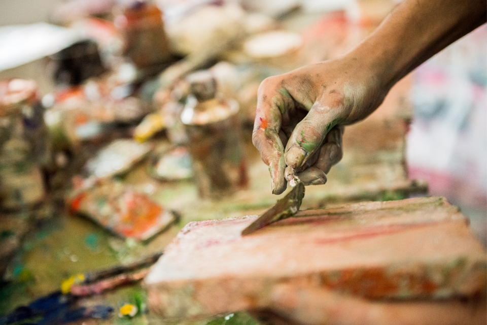 hand covered in paint holding a palette knife to put pink paint on a small canvas