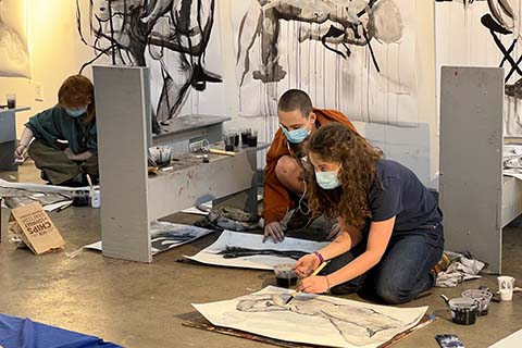 three students working on the ground with their art pieces with larger drawings hung up on the wall behind them