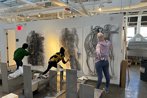 Rear view of 3 students working on large drawings on the wall of humanlike figures