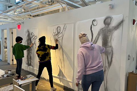 group of three students drawing humanlike figures on a large paper on the wall