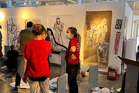 two students standing in the middle of an art room talking, with human figure drawings in the background