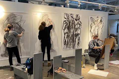 Three students in an art classroom with drawings hung up on the wall. Two students are drawing standing up while one student is sitting down drawing