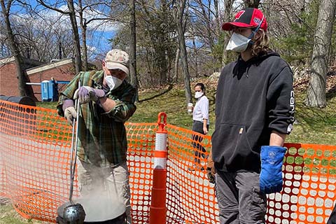 two individuals standing in front of a smoking raku can, removing a pottery item with tongs