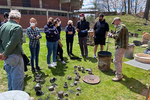 Group of students and instructors talking in front of rakued pieces. Christopher Frost is removing an item from the raku can
