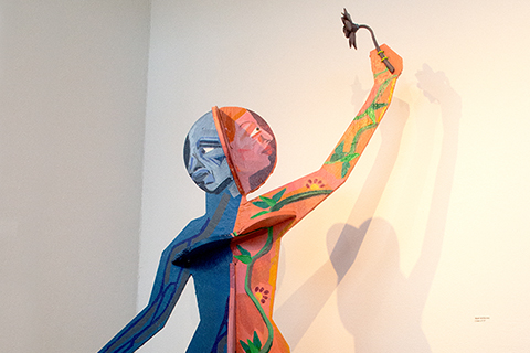 A five-foot tall by three-foot wide freestanding figurative wood sculpture. The figure is a split-faced and bodied person. One side is painted in warm colors with a smiling female looking upward at a 3D printed flower she is holding. The other side is painted in cool colors with a distraught male looking downward at his empty hand.