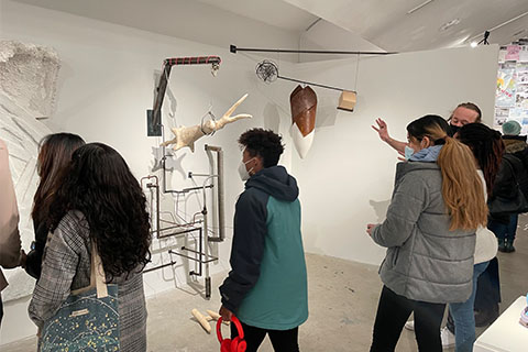 Students and guests standing in front of complex sculpture attached to the wall and hanging from the ceiling. One sculpture is a metal hook holding an appearing branch with metal poles below. The sculpture hanging from the ceiling is a lever made out of metal with a large metal object hanging from the middle.
