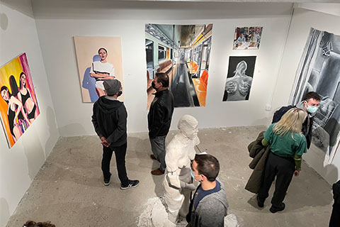 Students and guests standing in a room with artwork on the wall and a human sculpture in the middle