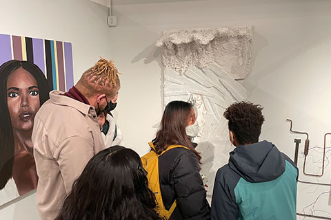 Group of people standing in front of artwork. Jonathan Joasil's piece on the right of a woman in front of stripes. Aeden Reid's piece on the right of a white sculpture.