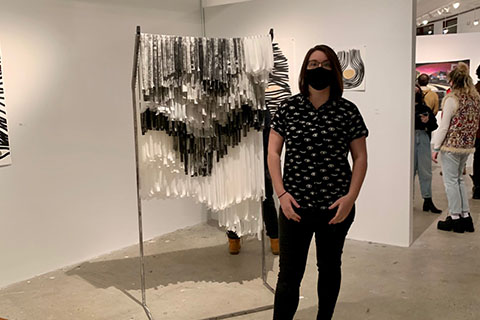 Kate Flake standing in front of a metal sculpture. White, gray, and black flimsy strips hanging from the metal poles