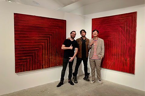 Lawler, Johnson and Yin posing in front of two large squares on a white wall. The squares are filled with lines swapping between red and dark red.