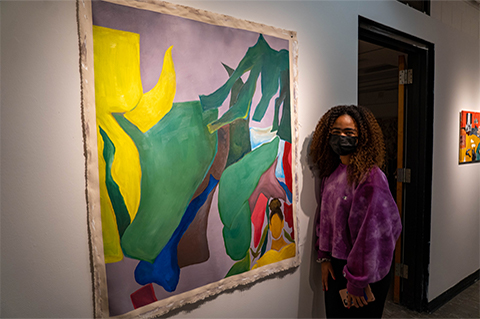Aria Smith standing in front of Smith's painting of colorful shapes on a canvas with an image of a woman in the bottom right