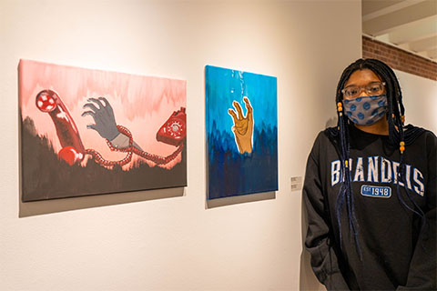 Bintou Baysmore standing in front of Baysmore's two paintings. The painting on the right is a hand reaching out of a blue abyss and the painting on the left is a hand caught up in a red telephone wire