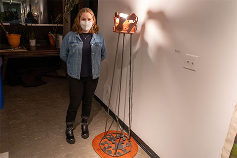 Hannah Rubin standing in front of Rubin's human size sculpture. Metal poles supporting wooden shapes that form a circle around a light bulb
