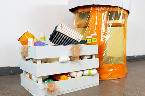 A tall empty orange pill bottle made of cardboard and see-through cellophane next to a crate filled with found items specifically homemade oversize stress balls, fidget cubes, and small pill bottles scattered throughout.