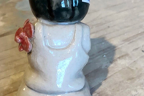 This 4.5 x 2.5 inch ceramic sculpture is of a seated figure with no feet or arms and whose head looks like a watermelon. On their left side where their arm would be there is a pink flower. 