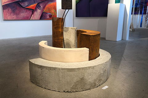 semicircular objects made out of concrete and metal are intertwined creating a sculpture in the center of a room with painting on the wall in the background