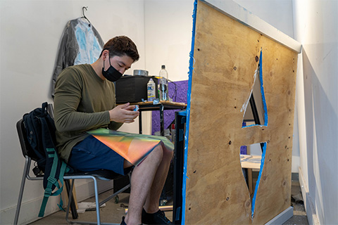 Wang sits in front of a large piece of wood with two triangle cut outs; Wang is working on this piece