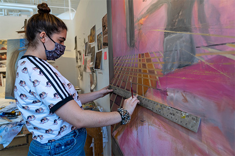 Angled view of Shire-Plumb holding a ruler up to an artwork on the wall; painting is of a pink hued room and a figure shape in gray