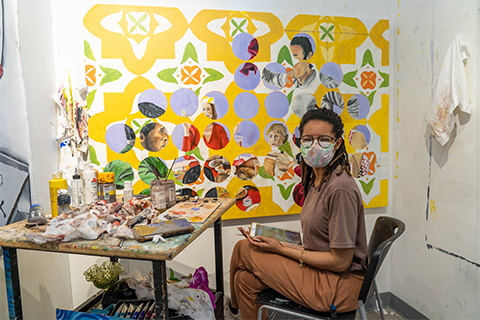 Brathwaite smiling for a photo with a mask on while sitting down in front of artwork that Brathwaite is working on; the painting is of a graphic pattern painted over portraits
