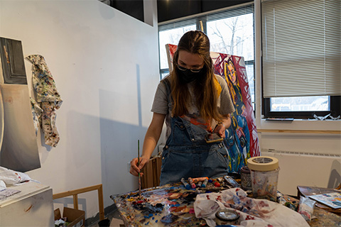 Young holding a phone in one hand and paint brush in the other dabbing paint; behind Young is a large canvas on an easel