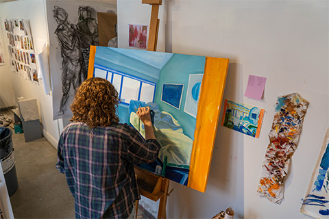 back view of donavan painting a blue pillow on an interior room painted in blue hues with an orange stripe on either side of the painting