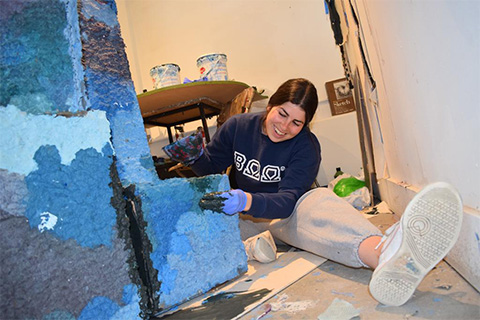 Fischer sitting on the floor and smiling while adding color to her blue-hued textured sculpture
