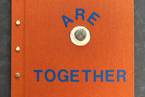An orange cloth-covered hardbound book with blue text reading, “We are together.” At the center of the cover beneath the word “are” is a perfectly round hole that shows a glimpse of the first page. The book is 13 inches tall, 10 3/4 inches wide, and 3/4 inches thick. 