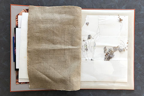 The back of Strauss’s page is a darker canvas which also shows through the pattern on the front of her page.  The eleventh page is artist Danielle Potwin. The form of this page is made by folding, ripping, and layering white paper. Drawn on the paper are squiggly black pen marks and a single verity of dried flowers collaged throughout. The pen peeks out from behind folded and layered paper where the flowers are sprinkled throughout. To preserve the page, it is bound by placing it in a sealed transparent pouch.