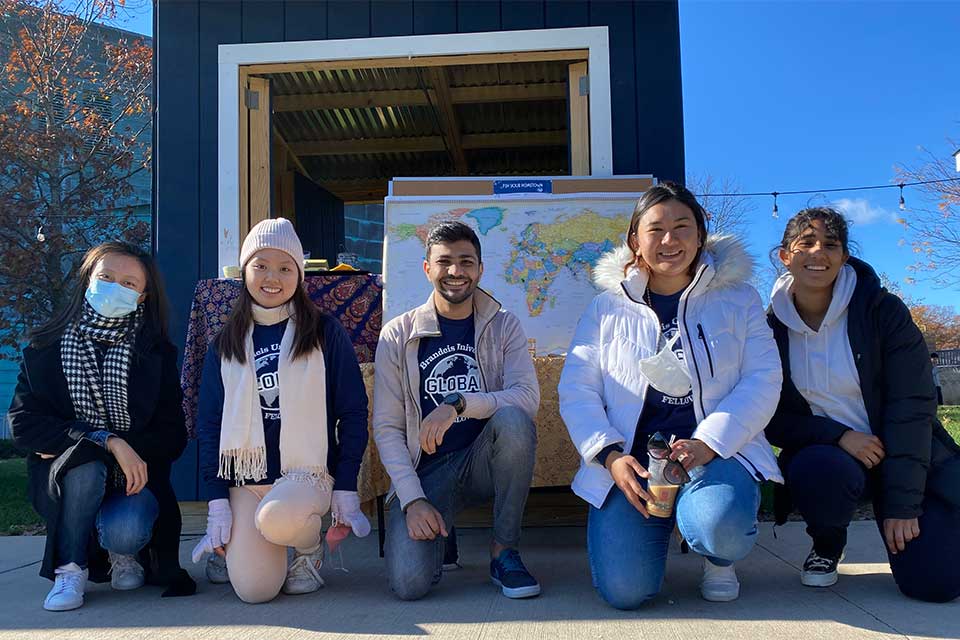 Five global fellows smiling outside in front of a world map on the Brandeis campus. 