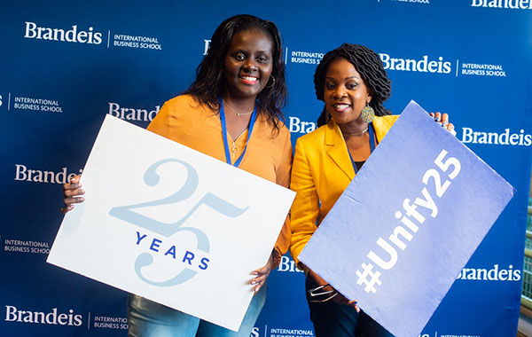 two female alumni in front of Brandeis International Business School banner holding Unify 25 signs