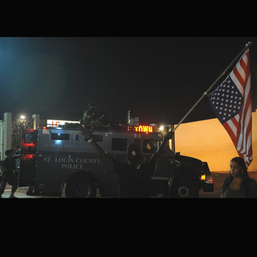 Black protesters wave an American flag at a police officer sitting in a heavy armored St. Louis police vehicle.