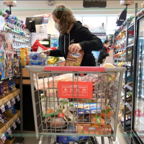 A woman in a mask with a full shopping cart in a supermarket aisle.