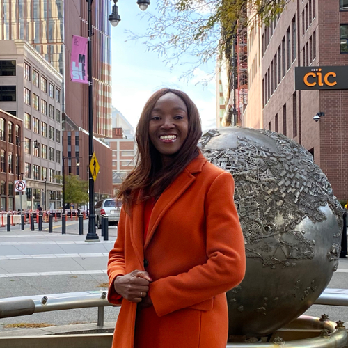 A portrait of Nova Diop in a burnt orange coat in Kendall Square, Cambridge.