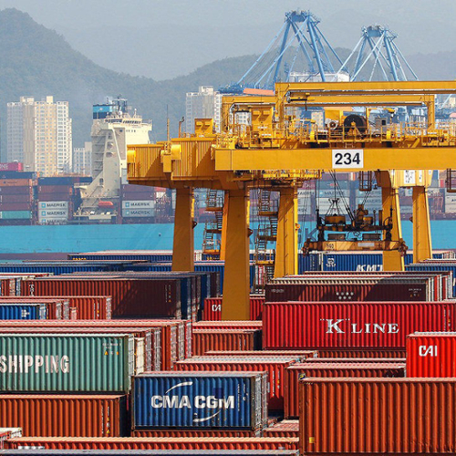 Stacks of shipping containers wait on a dock near a giant crane.