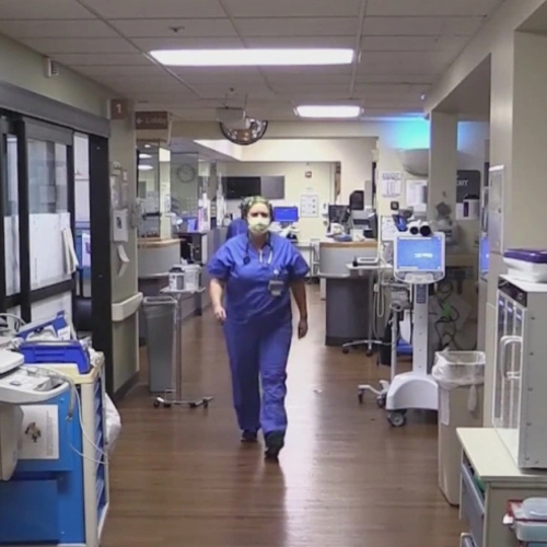 A nurse in blue scrubs and a face mask walks along a hall in a hospital.