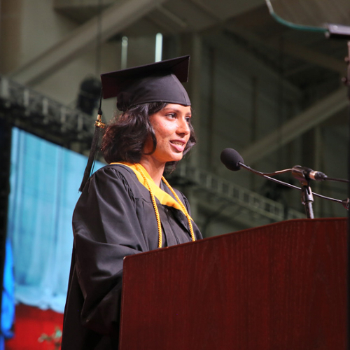 Apthi Harish in her cap and gown speaking at a podium during commencement.