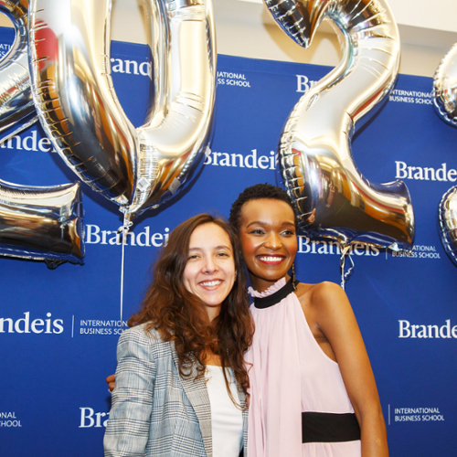 Two students pose in front of a Brandeis backdrop with gold 2023 balloons.