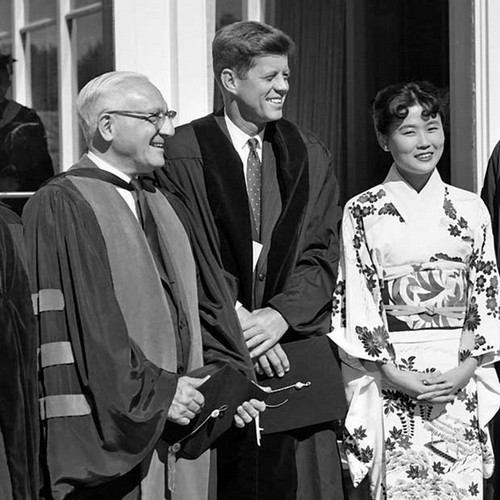 A black and white photograph of President John F. Kennedy in academic robes standing with Abrahm Sachar in robes and Wakako Kimoto Hironaka, MA’64 in a formal white floral kimono.