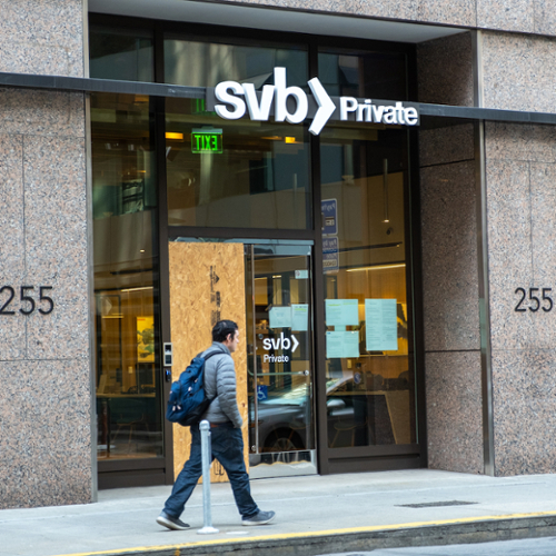 A man walks past a closed branch of Silicon Valley Bank, a glass-facade building with a large piece of plywood covering one door and the initials SVB above the door.