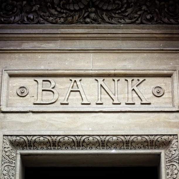 A close shot of a white marble building facade with the word "Bank" in block lettering above a door.