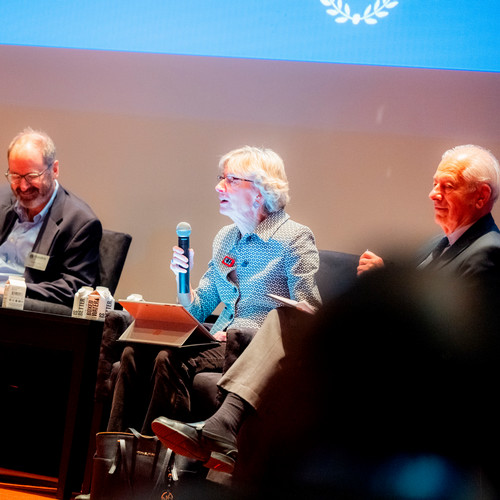 Dr. Catherine Mann speaks into a microphone while seated on stage next to Prof. George Hall and Prof. Peter Petri.