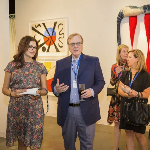 Paul Allen speaking to a small group of people with a collection of art hanging on the wall behind him.