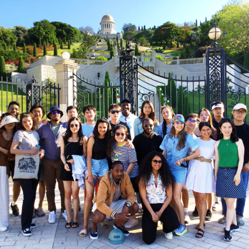 Two dozen students pose in front of a black metal gate in front of the Baha'i Gardens, which rise up the slope behind them to a building with a gold-domed cupola.