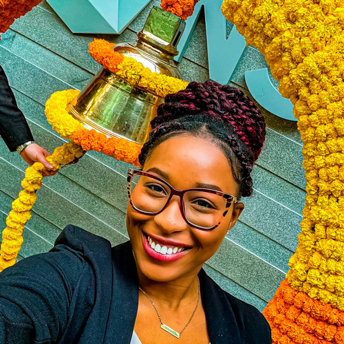 Djounia Saint-Fleurant smiles for a selfie with a ring of yellow and orange flowers and a gold bell behind her.