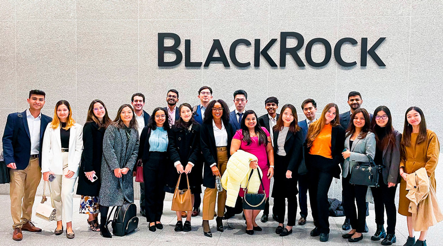 A group of students in business dress pose in front of a white marble wall with the name "BlackRock" behind them.