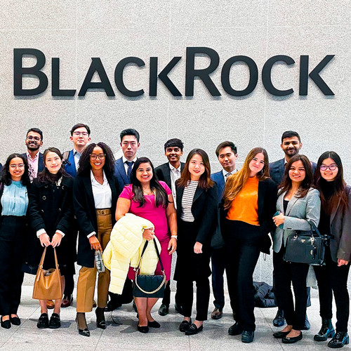 A group of students in business dress pose in front of a white marble wall with the name "BlackRock" behind them.