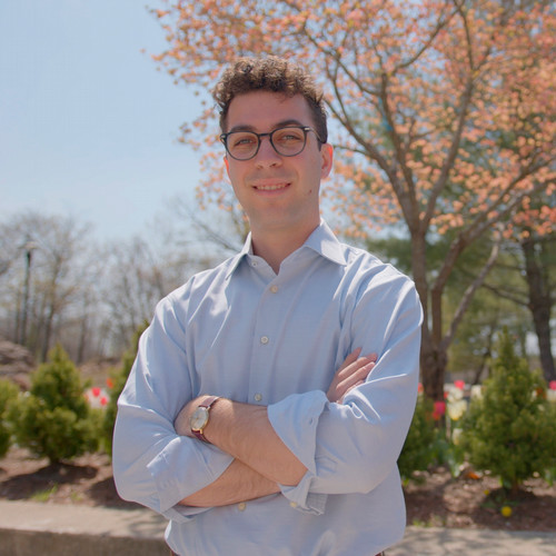 Jaden Menzer smiles with his arms crossed while standing outside in front of a blossoming tree.