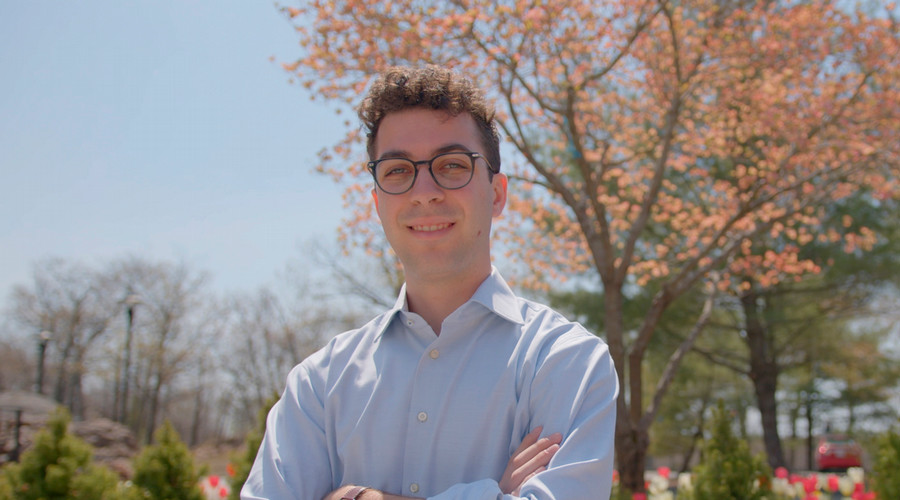 Jaden Menzer smiles with his arms crossed while standing outside in front of a blossoming tree.