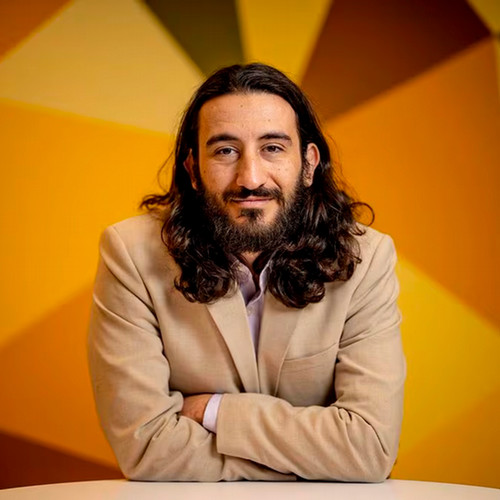 Eric Moyal, with a beard and long black hair in a tan suit smiles for the picture while leaning forward on a table.